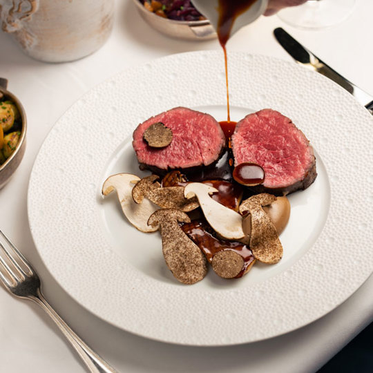 Sauce being poured over a plate of beef and mushrooms that is on a laid table